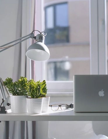 A desk in front of a window with a laptop, succulents, and lamp on top of it.